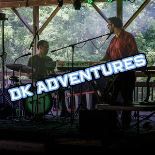 Dom and Kevin of DK Adventures performing on an outdoor wooden stage surrounded by trees — one on drums, one on keyboard and vocals — with a "DK Adventures" text overlay.