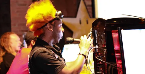 Mars Magma performing live in a bright orange wig and headlamp behind a bank of game consoles and a monitor, with the "Hipscore: The Happy Hour for Gamers" logo overlaid.