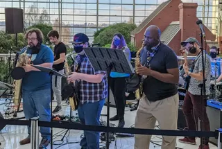 Quick Save performing live outdoors, members playing guitar and singing on a stage against a city skyline backdrop.